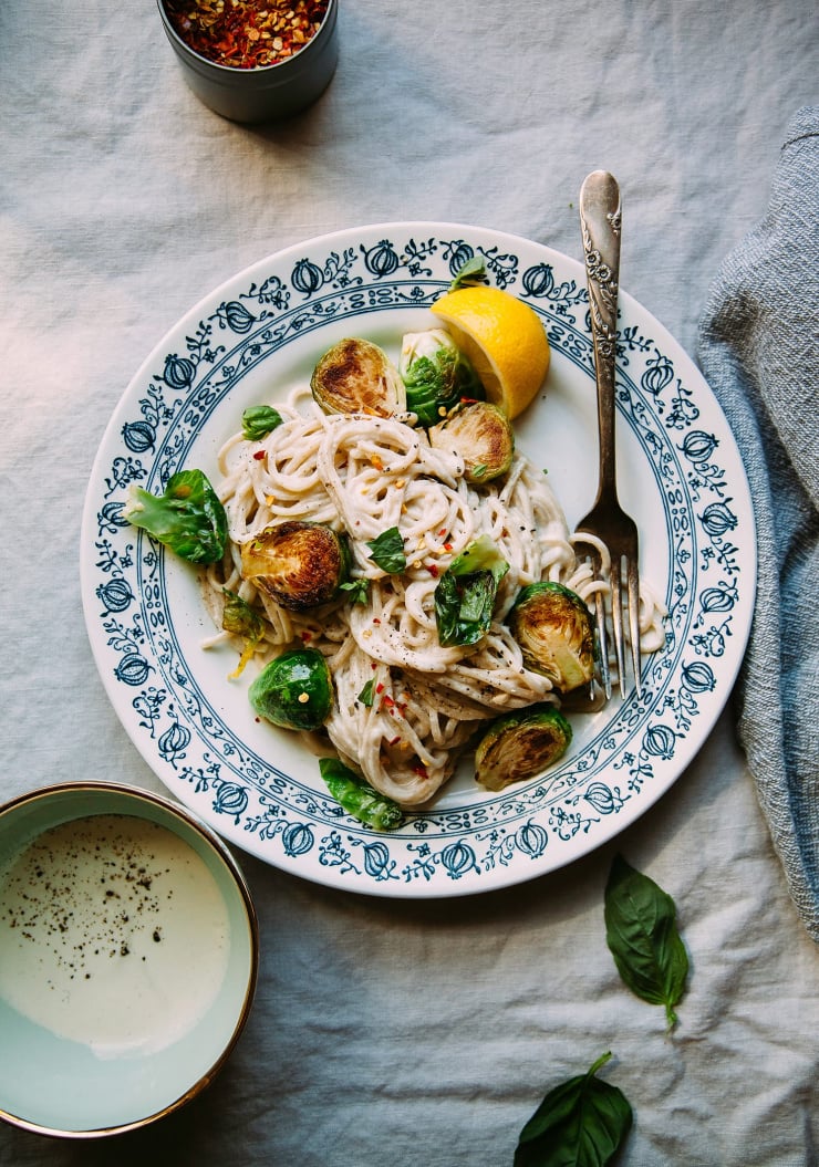 An overhead shot of creamy miso pasta with Brussels sprouts. The plate it is served on is white with a blue detail around the edge. The photo is shot on a beige linen tablecloth.