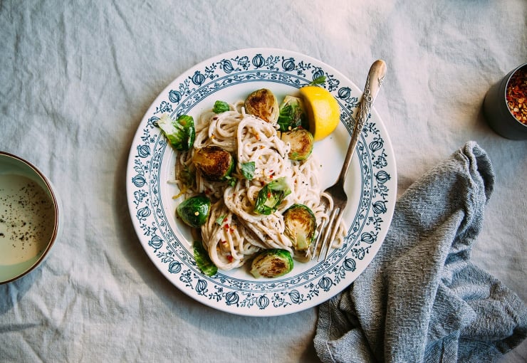 An overhead shot of creamy miso pasta with Brussels sprouts. The plate it is served on is white with a blue detail around the edge. The photo is shot on a beige linen tablecloth.