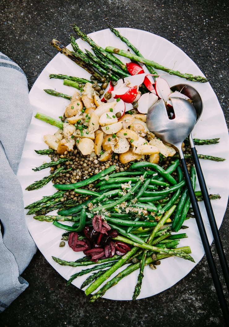 An overhead shot of a vegan lentil niçoise-inspired salad with potatoes, green beans, and asparagus. The white platter of salad is on a dark grey background.