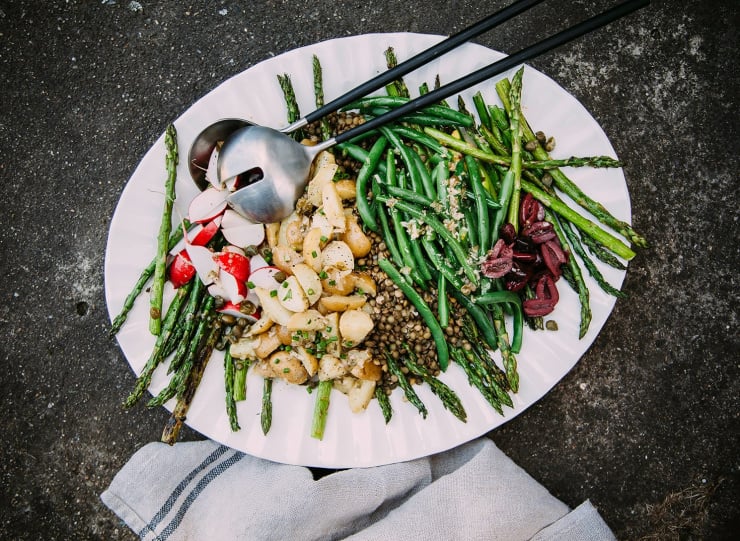 An overhead shot of a vegan lentil niçoise-inspired salad with potatoes, green beans, and asparagus. The white platter of salad is on a dark grey background.