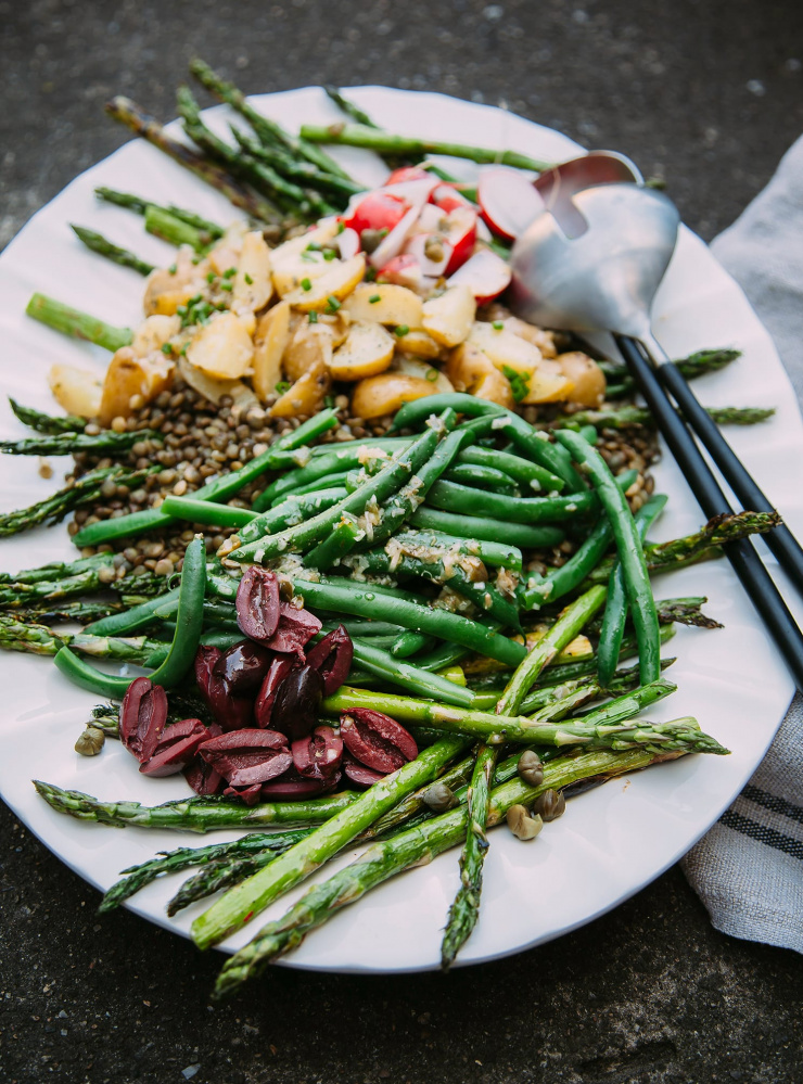 A 3/4 angle, up close shot of a vegan lentil niçoise-inspired salad with potatoes, green beans, and asparagus. The white platter of salad is on a dark grey background.