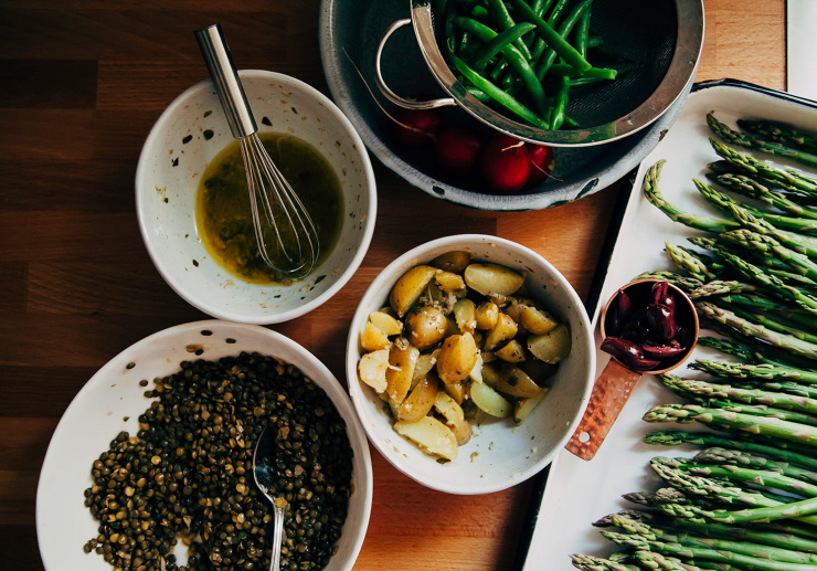 An overhead shot of prepped components in bowls for a composed salad. Included here: cooked green beans, asparagus, olives, cooked potatoes, lentils, and a bowl of vinaigrette.