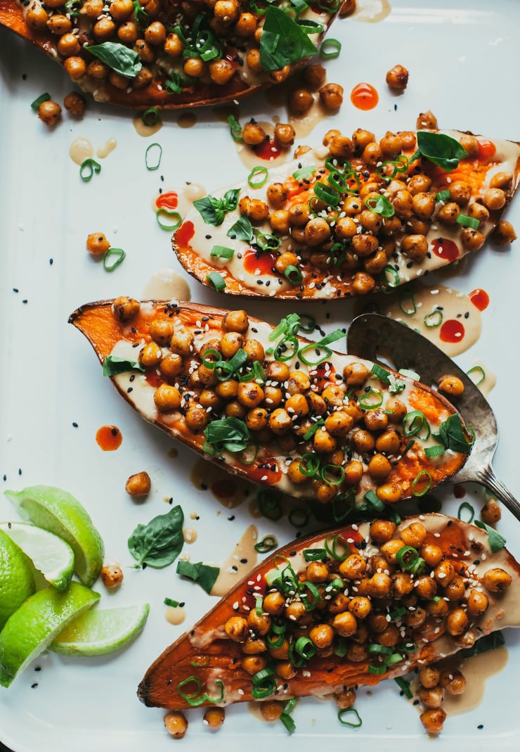 An overhead shot of roasted sweet potato halves stuffed with spiced chickpeas and chopped herbs. A tahini sauce is drizzled over the top and lime wedges are served to the side.