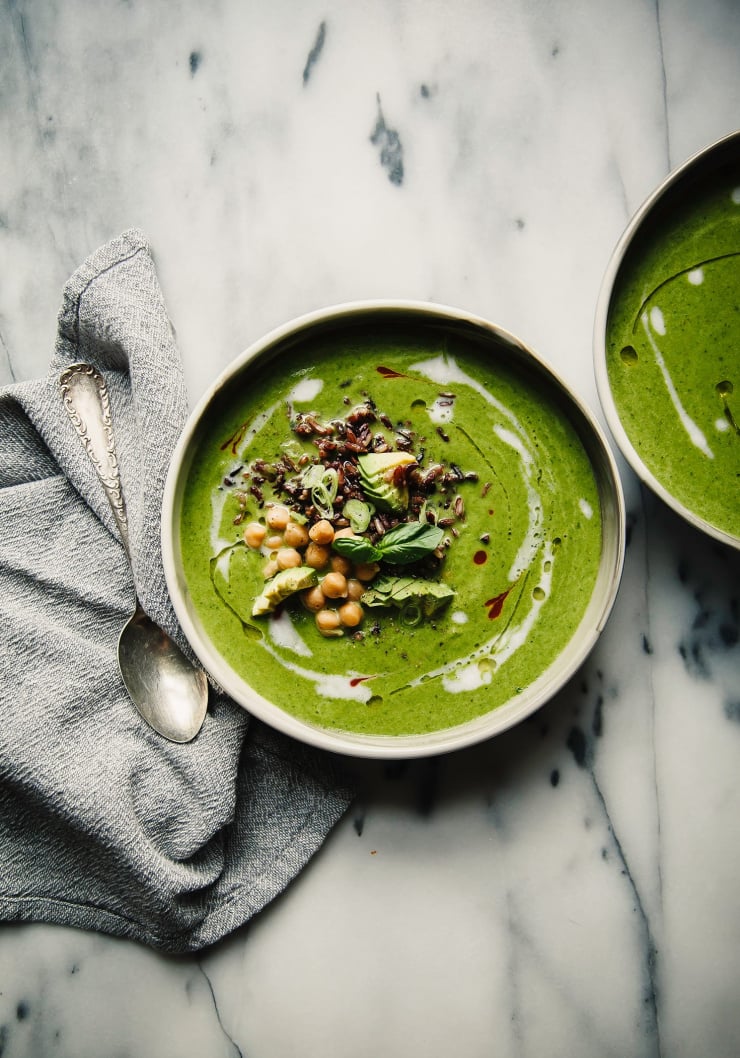 An overhead shot of bright green coconut green soup on top of a white marble background. There is a grey napkin nearby. The soup is garnished with cooked black rice, chickpeas, avocado, and fresh herbs.