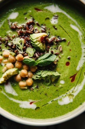 An up close, overhead shot of coconut green soup in a wide bowl. The bowl is topped with black rice, chickpeas, diced avocado, and fresh basil. The bright green soup is streaked with coconut milk and hot sauce.