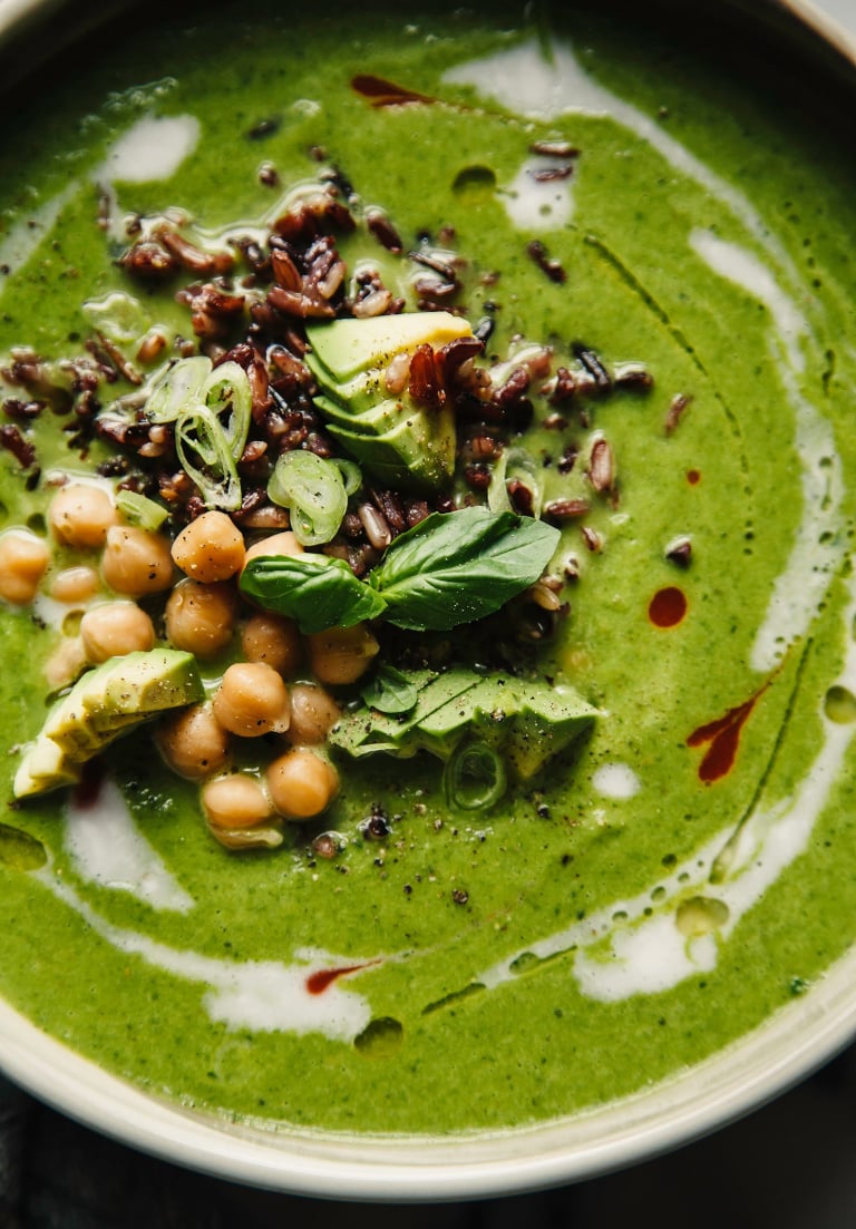 An up close, overhead shot of coconut green soup in a wide bowl. The bowl is topped with black rice, chickpeas, diced avocado, and fresh basil. The bright green soup is streaked with coconut milk and hot sauce.