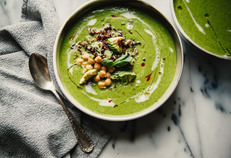 An overhead shot of bright green coconut green soup on top of a white marble background. There is a grey napkin nearby. The soup is garnished with cooked black rice, chickpeas, avocado, and fresh herbs.