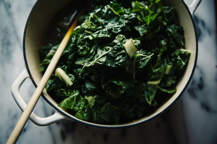 A white Dutch oven with tons of chopped chard added to it. A wooden spoon is sticking out of the pot.