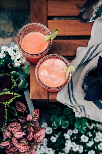 An overhead shot of two glasses of pink summer bliss juice on a wood table with plants to the side. Both glasses are garnished with lime wheels.