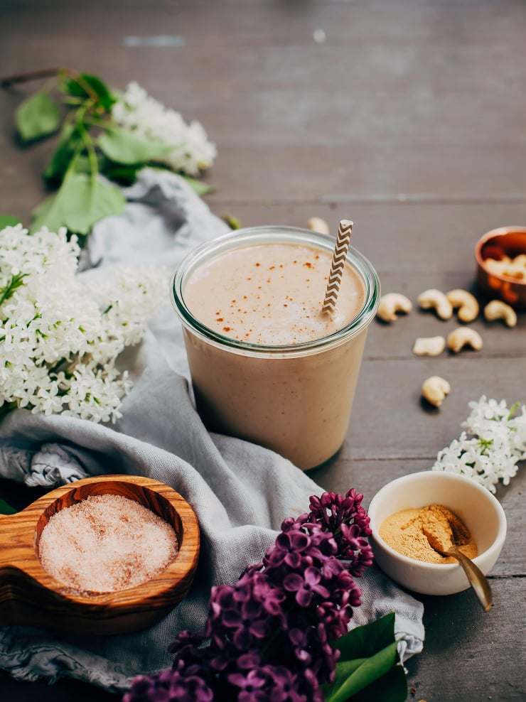A head on shot of a maca caramel frap with some lilac flowers, a pinch bowl of salt, and raw cashews nearby.