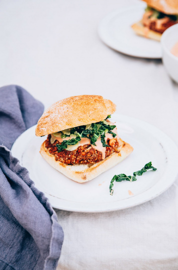 A slight 3/4 angle shot of a bloody mary veggie burger topped with a creamy sauce and some shredded kale on a white plate with a dark blue napkin nearby.