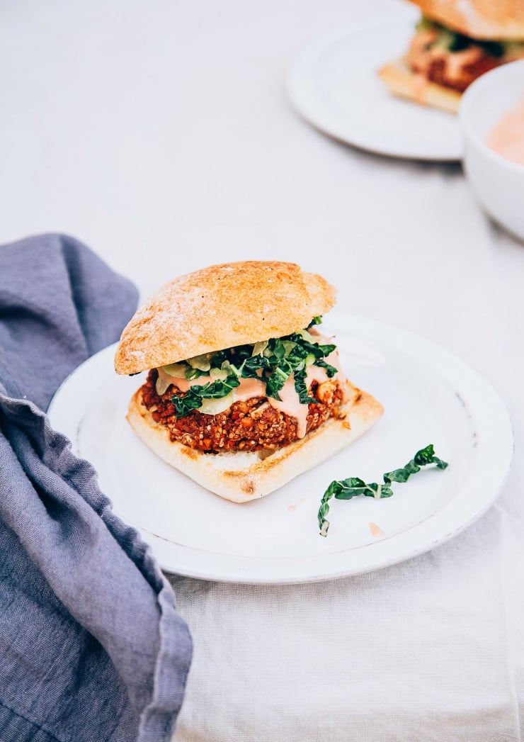 A slight 3/4 angle shot of a bloody mary veggie burger topped with a creamy sauce and some shredded kale on a white plate with a dark blue napkin nearby. Part of a roundup of vegan dinner recipes.