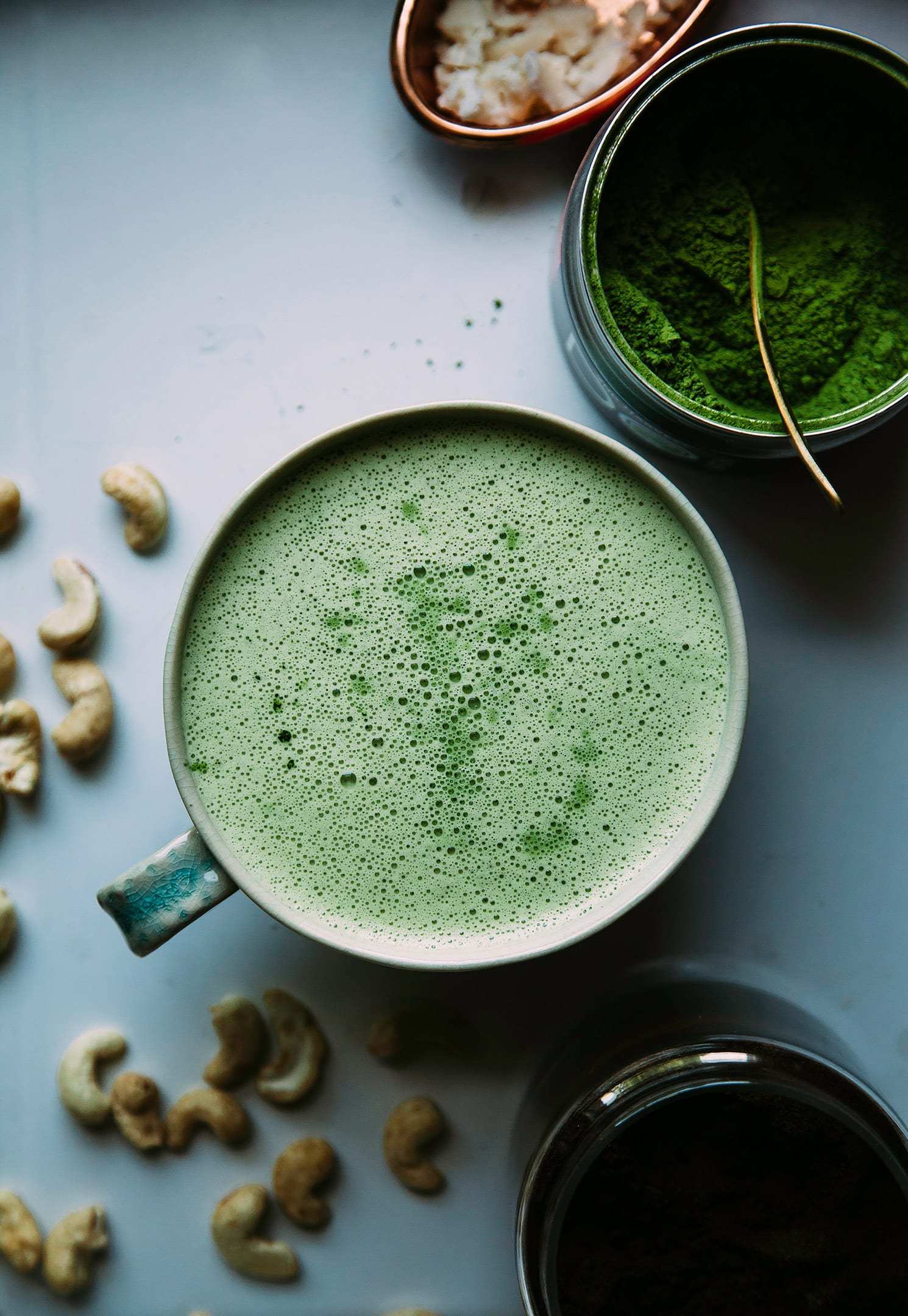 An overhead shot of a mellow green, coconut cashew matcha latte with a bowl of matcha nearby and some cashews strewn about.