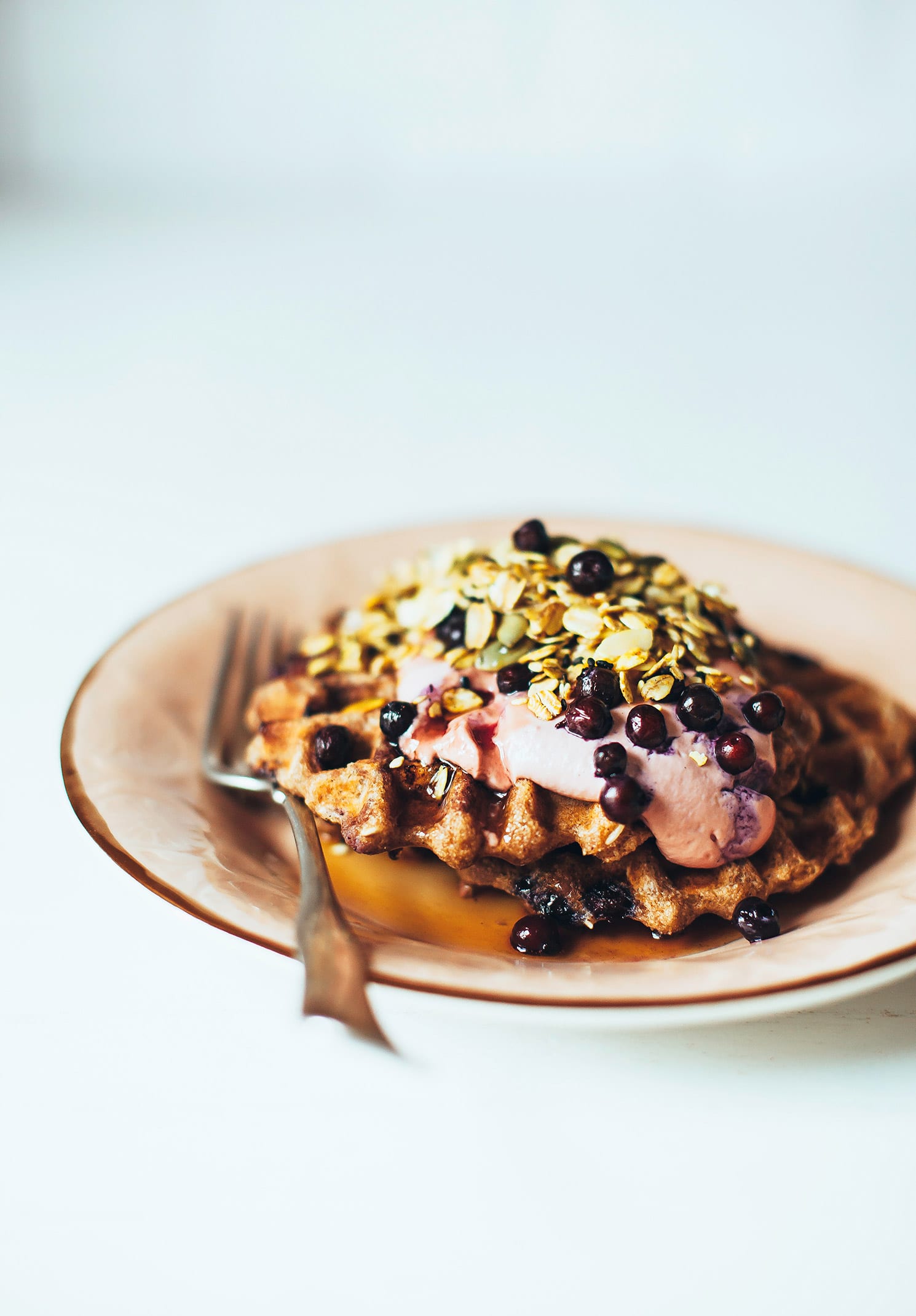 A head-on shot of blueberry spelt waffles topped with a light pink cream, blueberries, and a golden yellow turmeric granola. The waffles are on a light pink plate rimmed with gold.