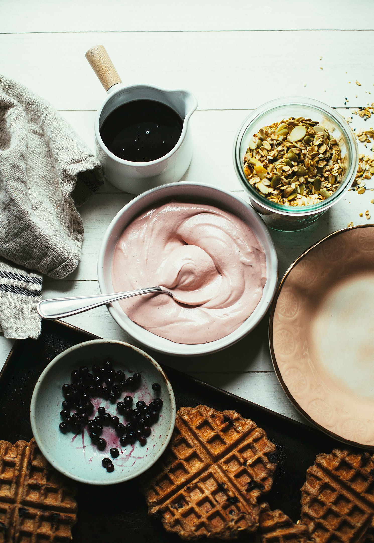 An overhead shot of a bowl of pink cream, a jar of golden yellow turmeric granola, a sheet pan of cooked waffles, a carafe of maple syrup, and a small bowl with some thawed frozen blueberries.