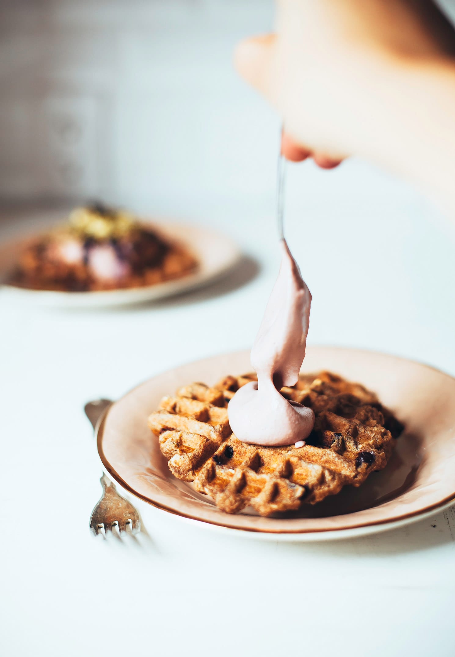 A head on image shows a hand using a spoon to dollop some thick pink cream on top of a cooked waffle.