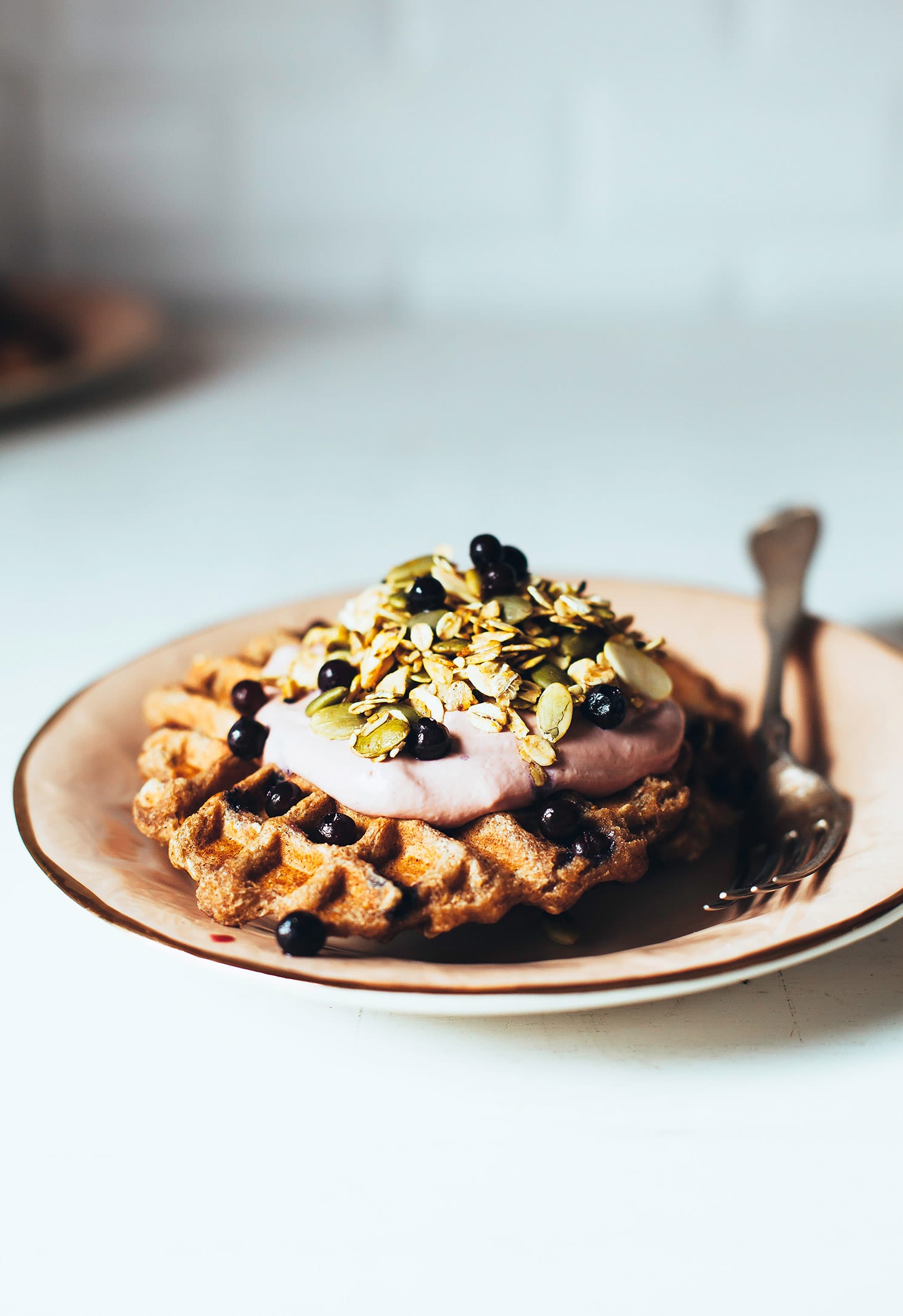 A head-on shot of blueberry spelt waffles topped with a light pink cream, blueberries, and a golden yellow turmeric granola. The waffles are on a light pink plate rimmed with gold.