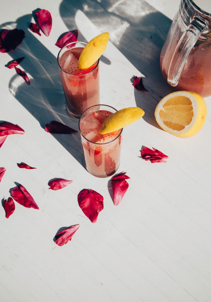An overhead shot of two glasses of strawberry rosewater chia fresca in direct sunlight. The drinks are garnished with lemon wedges and surrounded by bright pink rose petals.