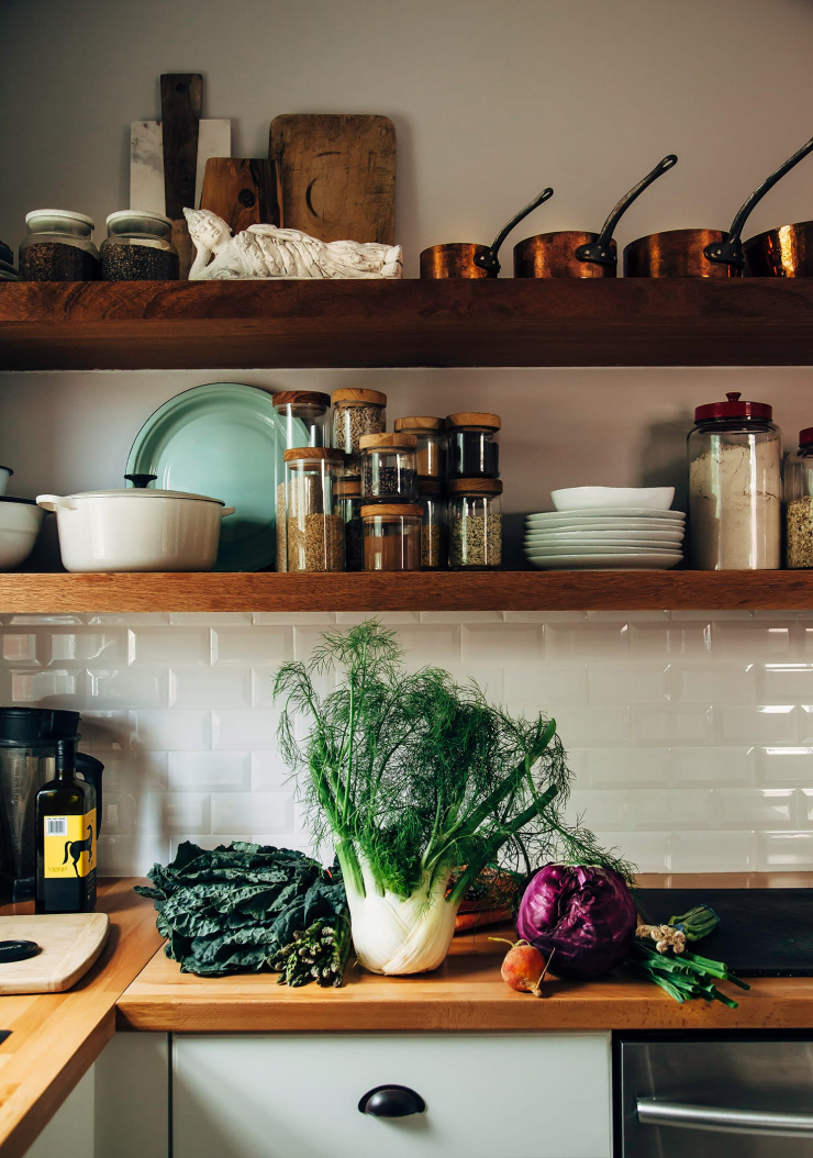A straight on shot of a kitchen scene with wooden open shelving, butcher block counters with vegetables on top, and dim natural lighting.