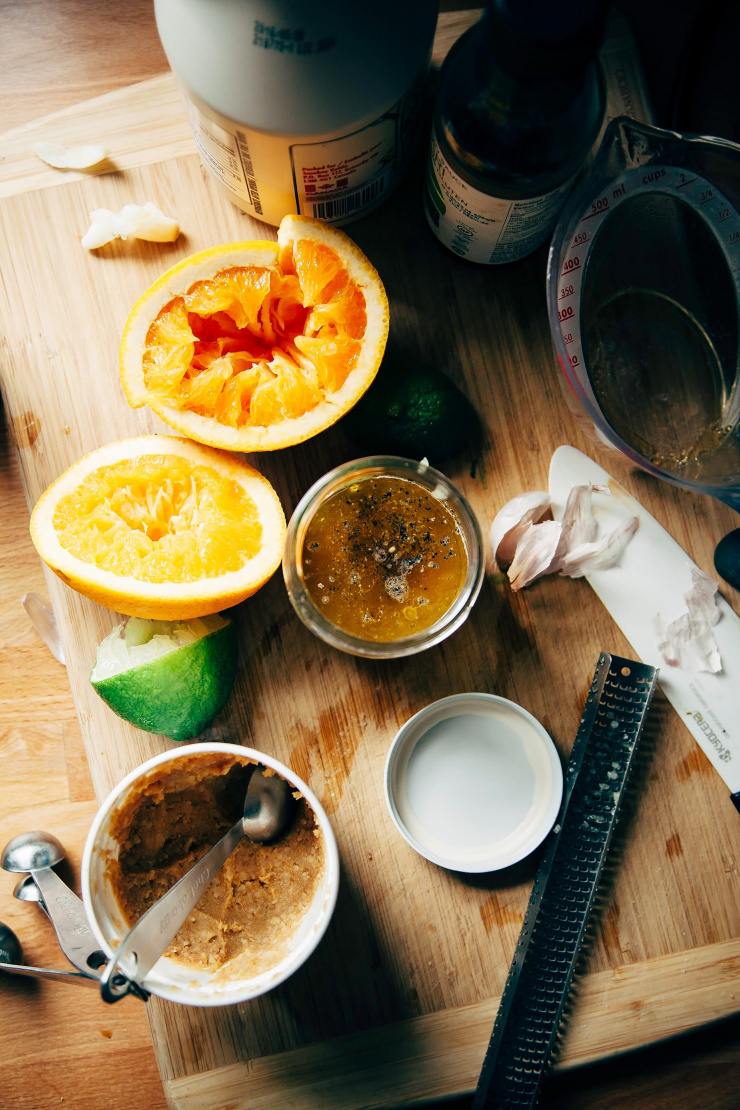 An overhead shot of ingredients for a salad dressing on a bamboo cutting board: orange halves, lime half, miso paste, garlic, tamari, and maple syrup.