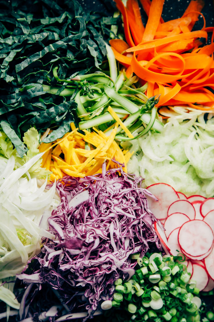 An overhead shot of prepped vegetables: red cabbage, fennel, green onions, radishes, golden beets, carrots, asparagus, celery, and kale all shaved thin or finely sliced.
