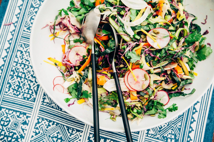 An overhead shot of a citrus miso slaw with 10 different types of shaved/thinly sliced vegetables; all in a white bowl with matte black/steel serving tongs.