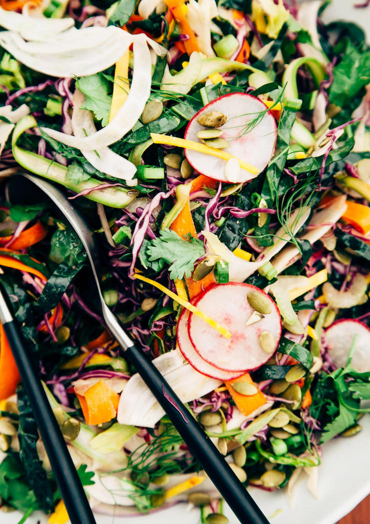 An overhead shot of a citrus miso slaw with 10 different types of shaved/thinly sliced vegetables; all in a white bowl with matte black/steel serving tongs.