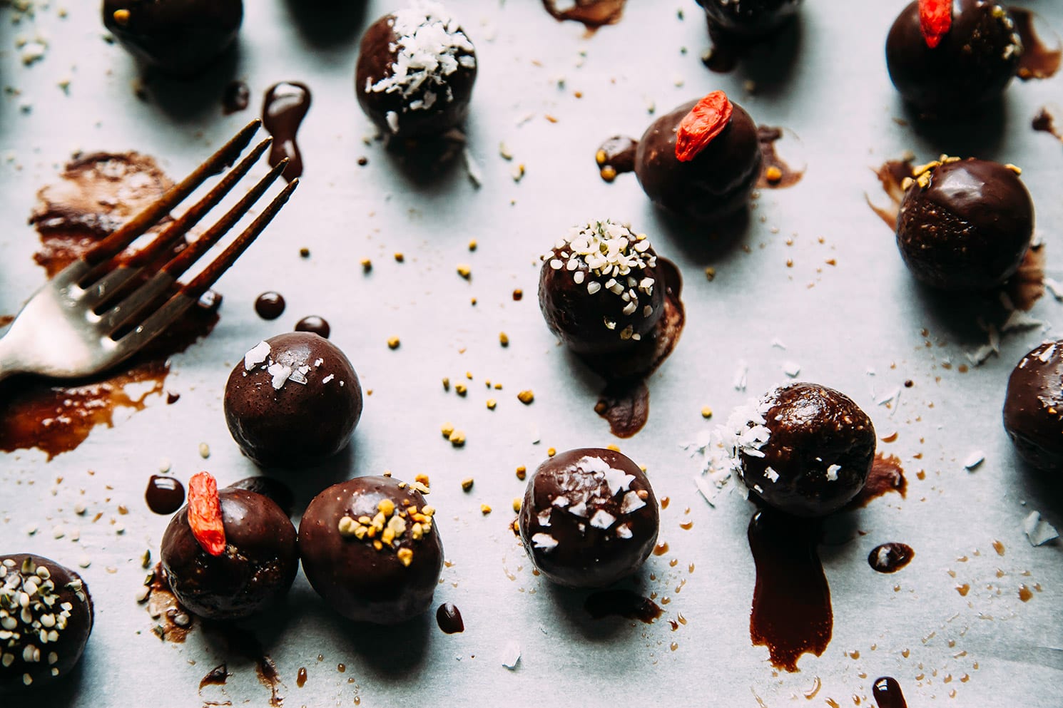 An overhead shot of rolled up superfood brownie bites that are dipped in melted chocolate. Each one is garnished with either bee pollen, goji berry or shredded coconut