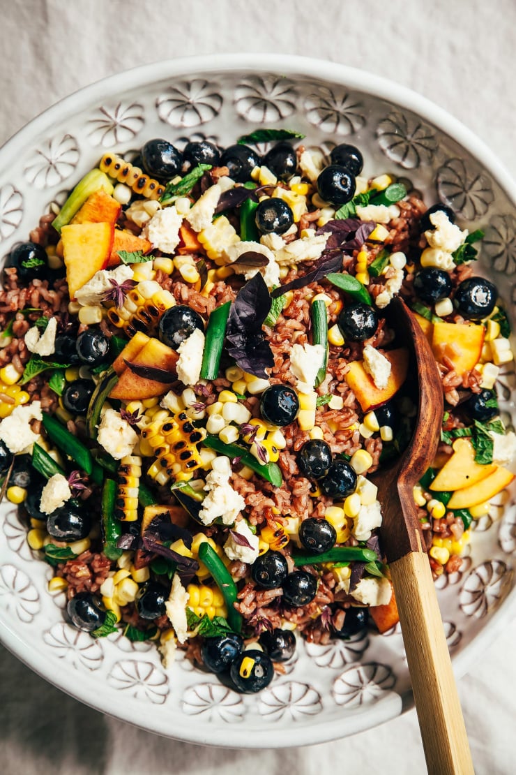 An up close, overhead shot of a summer picnic salad with corn, blueberries, garlic scapes, pink rice, and pieces of vegan "feta" cheese.