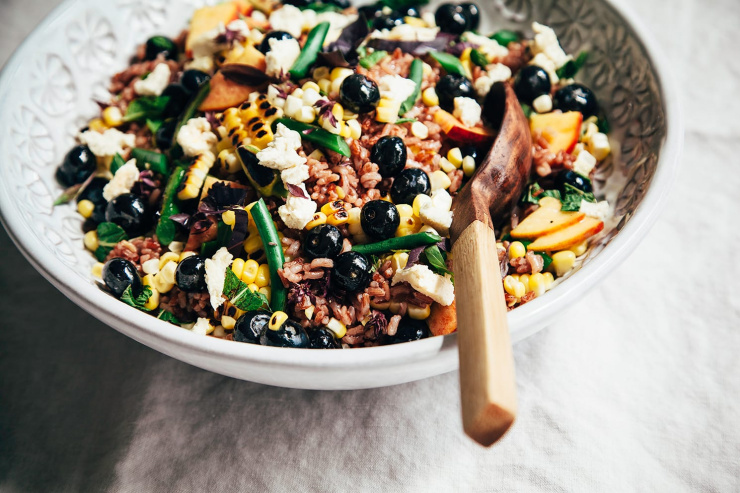 A 3/4 angle shot of a summer picnic salad with corn, blueberries, garlic scapes, pink rice, and pieces of vegan "feta" cheese.