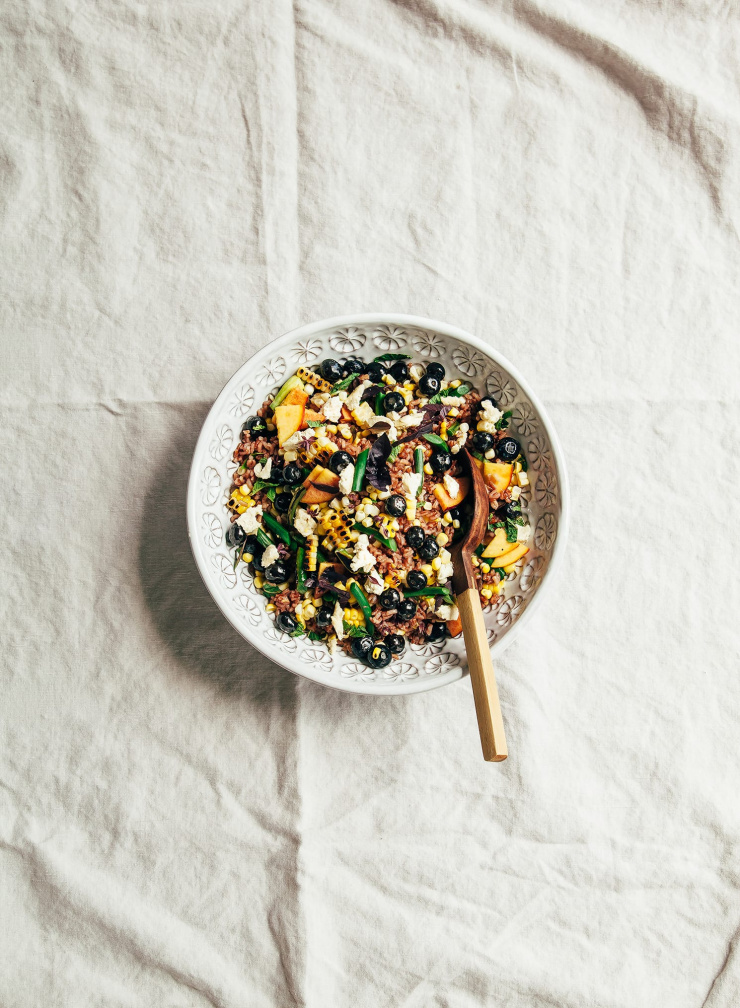 A zoomed out overhead shot of a summer picnic salad with corn, blueberries, garlic scapes, pink rice, and pieces of vegan "feta" cheese.