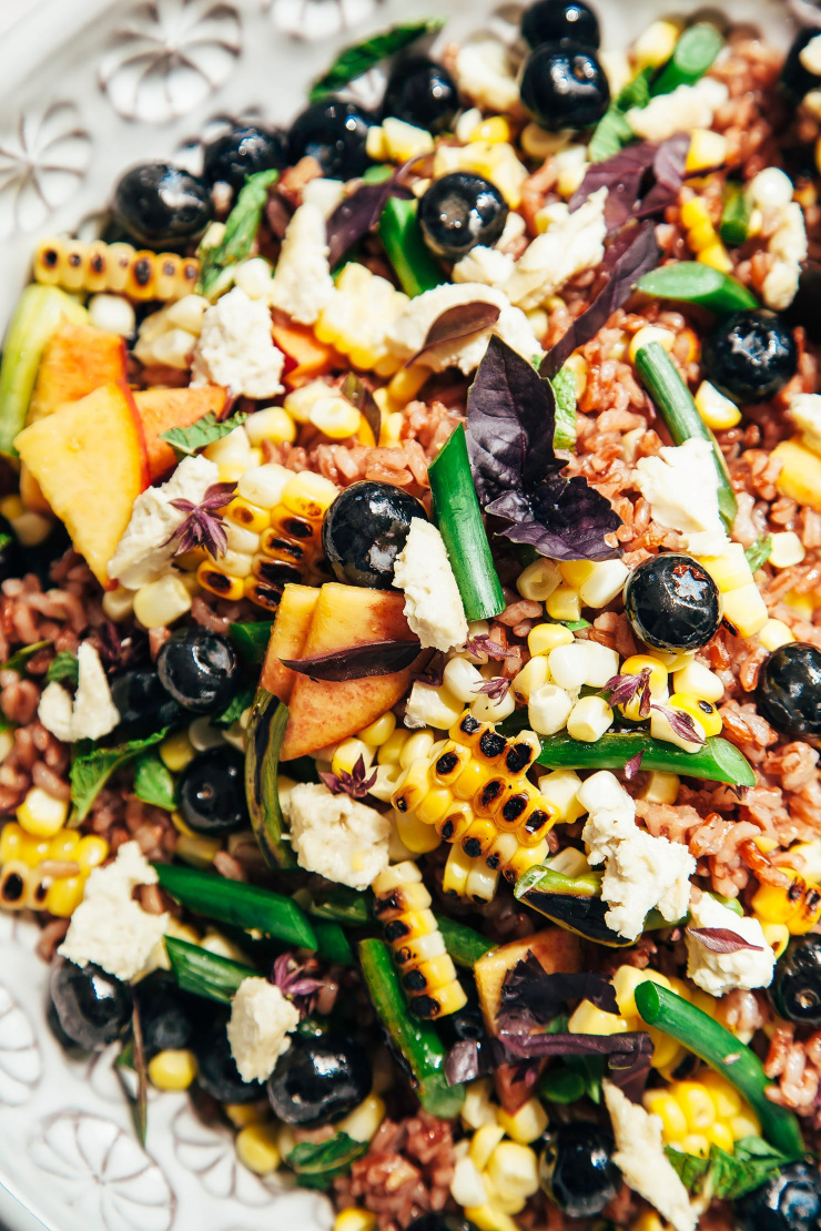 An up close, overhead shot of a summer picnic salad with corn, blueberries, garlic scapes, pink rice, and pieces of vegan "feta" cheese.