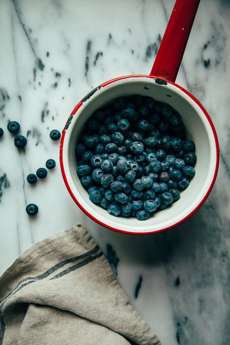 An overhead shot of fresh blueberries in a red-handled enamelware pot on a marble background.