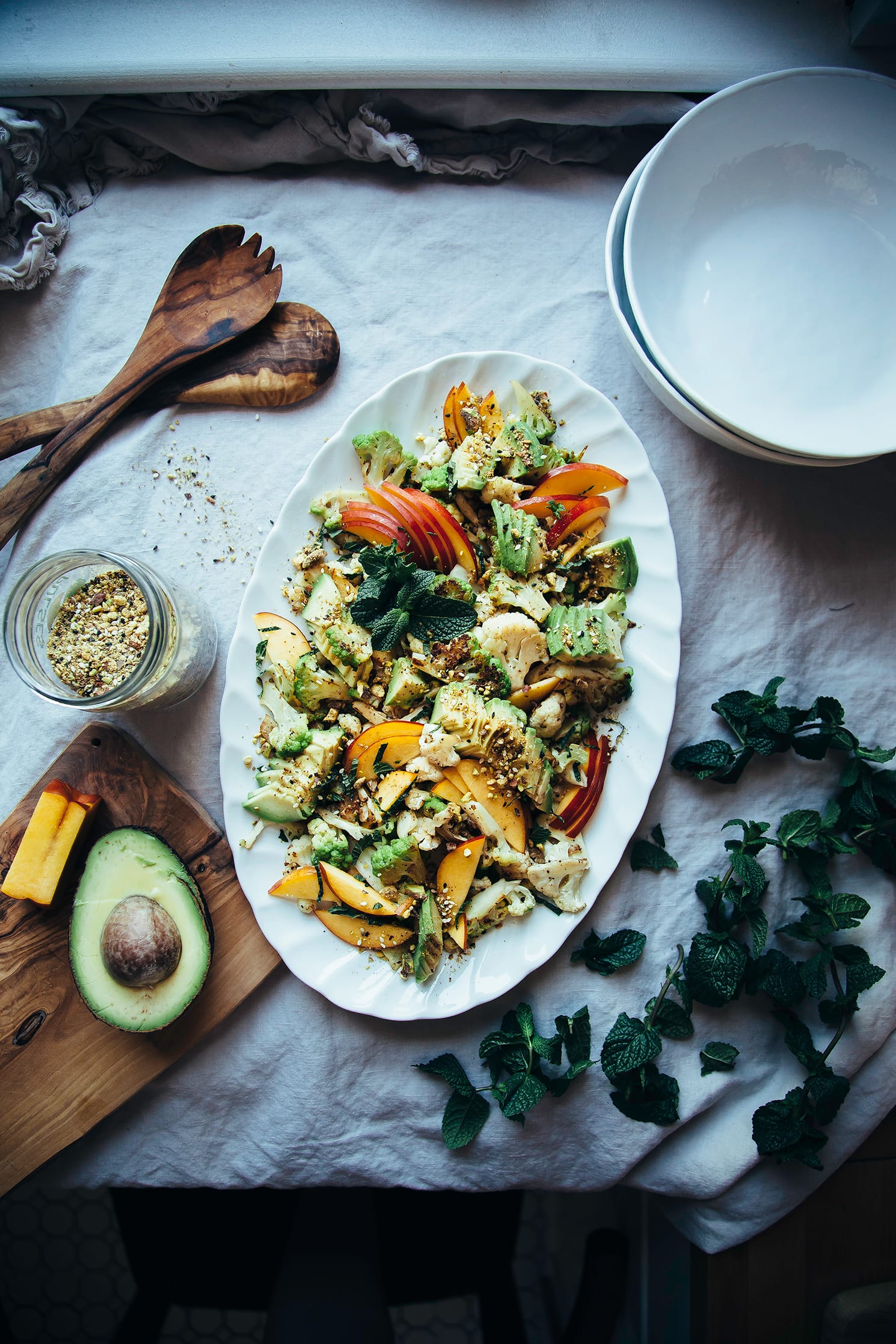 An overhead shot of a cauliflower salad on a white oval platter, topped with sliced nectarines, avocado, fresh mint leaves, and a nut/seed spice mixture.