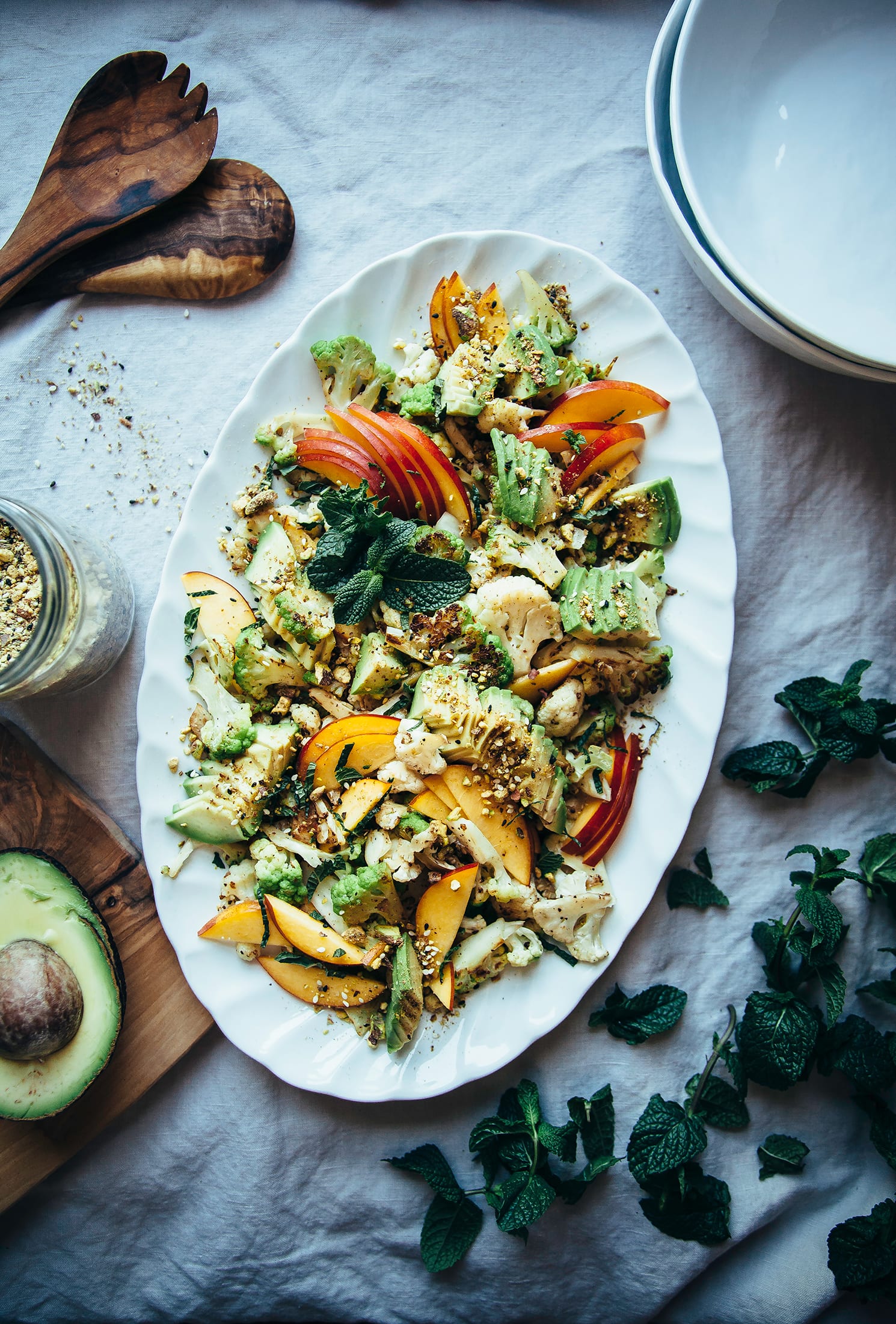 An overhead shot of a cauliflower salad on a white oval platter, topped with sliced nectarines, avocado, fresh mint leaves, and a nut/seed spice mixture.
