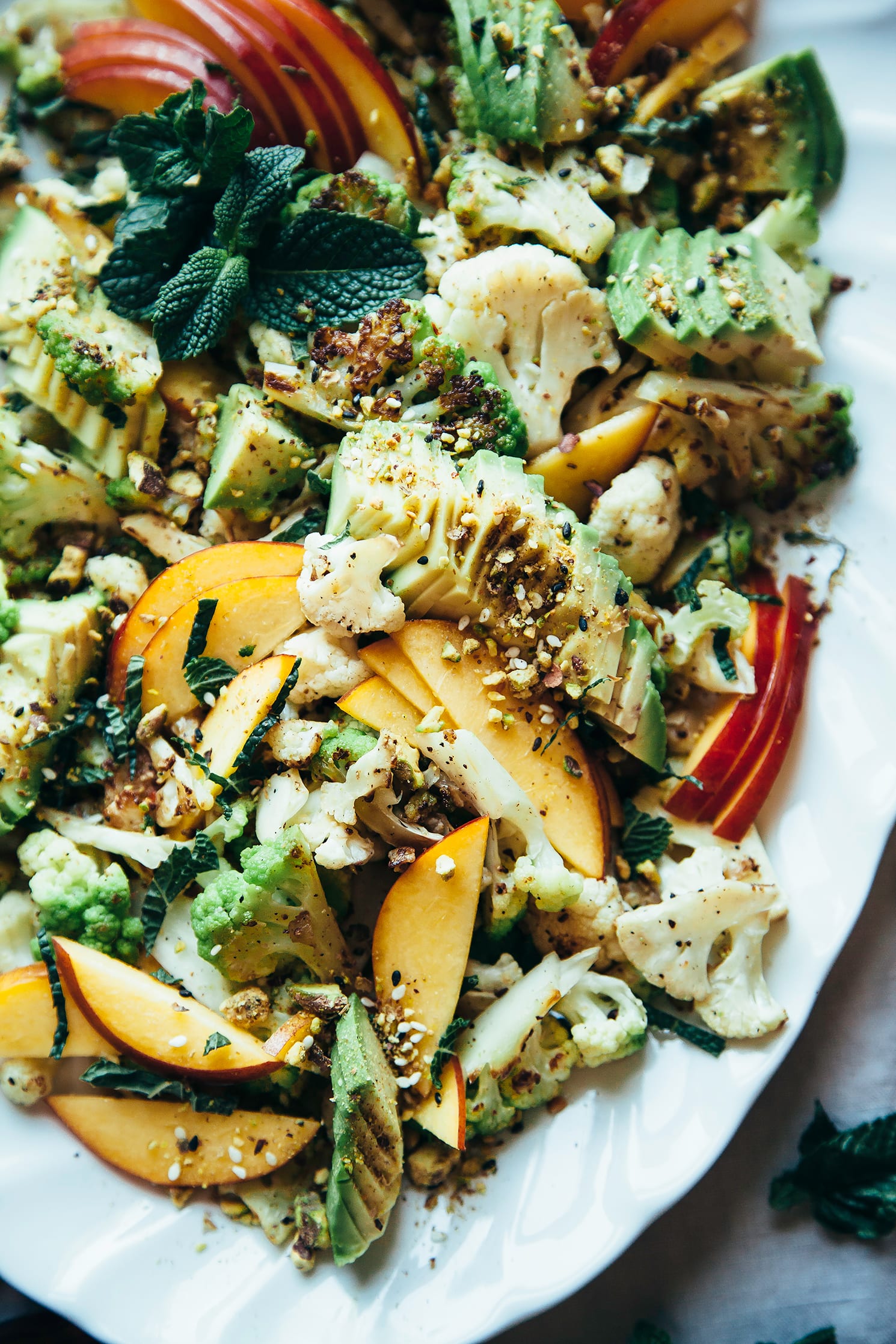 An overhead shot of a cauliflower salad on a white platter, topped with sliced nectarines, avocado, fresh mint leaves, and a nut/seed spice mixture.