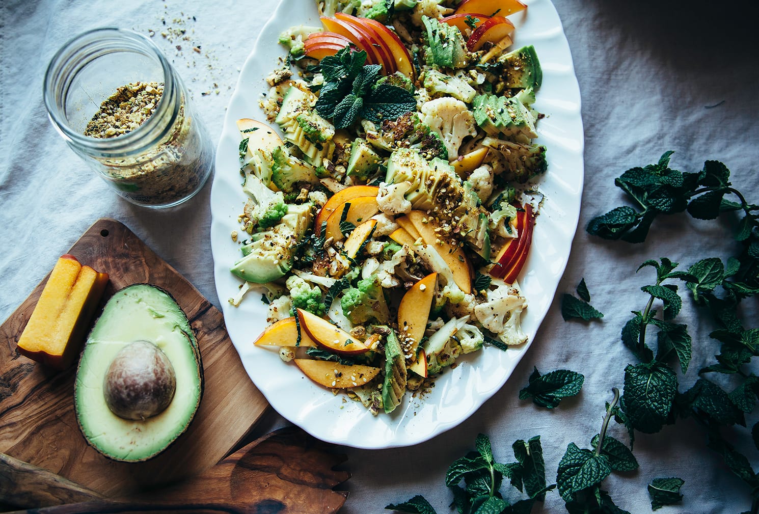 An overhead shot of a cauliflower salad on a white oval platter, topped with sliced nectarines, avocado, fresh mint leaves, and a nut/seed spice mixture.