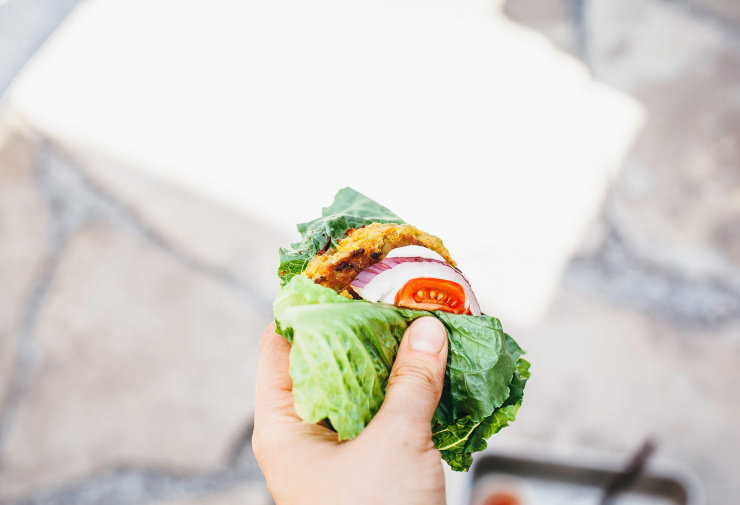 An overhead shot of a hand holding a lettuce-wrapped golden split pea patty with tomato, red onion and mayo inside.