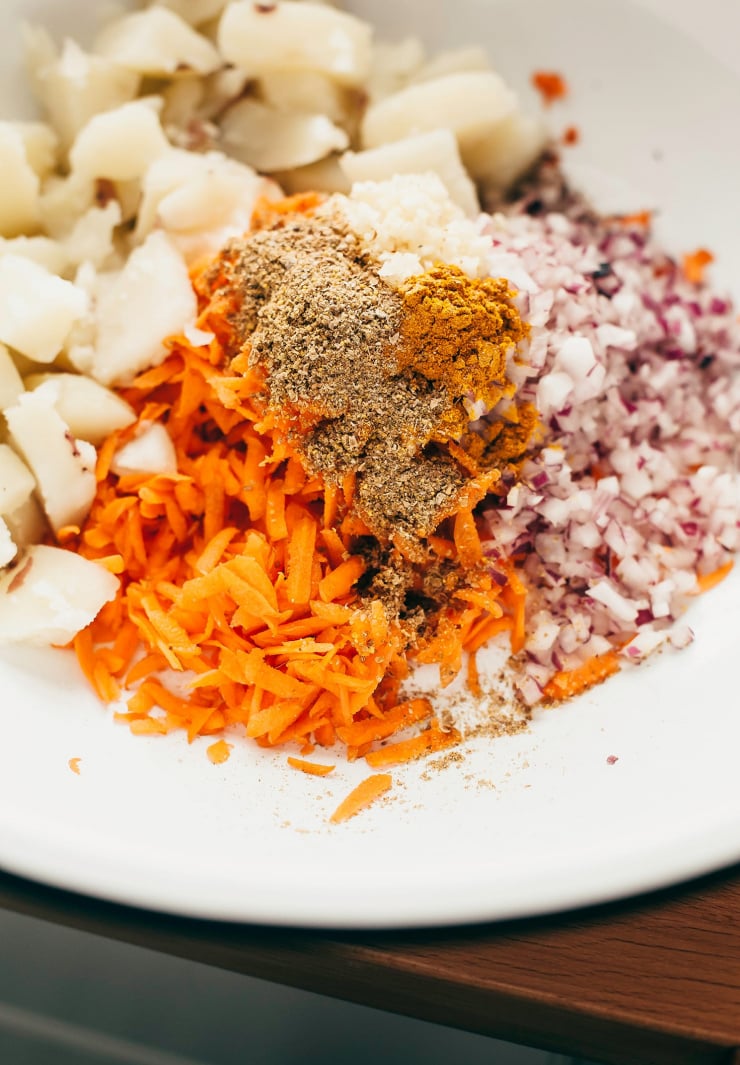 An overhead shot of cooked potatoes and carrots in a white bowl with some diced onion and spices.