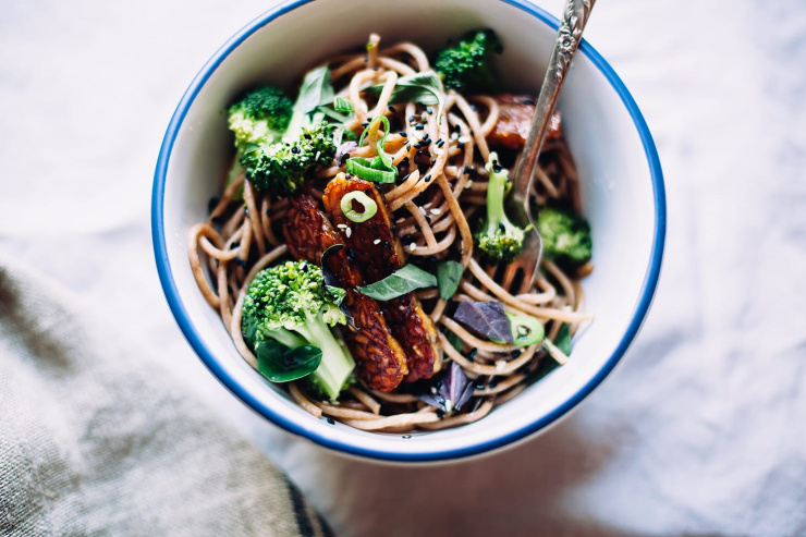 An overhead shot of a bowl of noodles with slices of tempeh and cooked broccoli pieces.