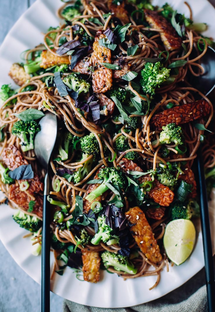 An overhead shot of noodles, broccoli, and seared tempeh slices on a pink platter, all coated in a deep brown, lightly textured sauce. Chopped basil is sprinkled on the top and a wedge of lime is seen to the side. Part of a roundup of vegan dinner recipes.