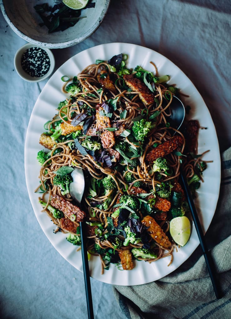 An overhead shot of garlic sesame noodles on a white platter. The dish also features broccoli florets and chopped herbs.