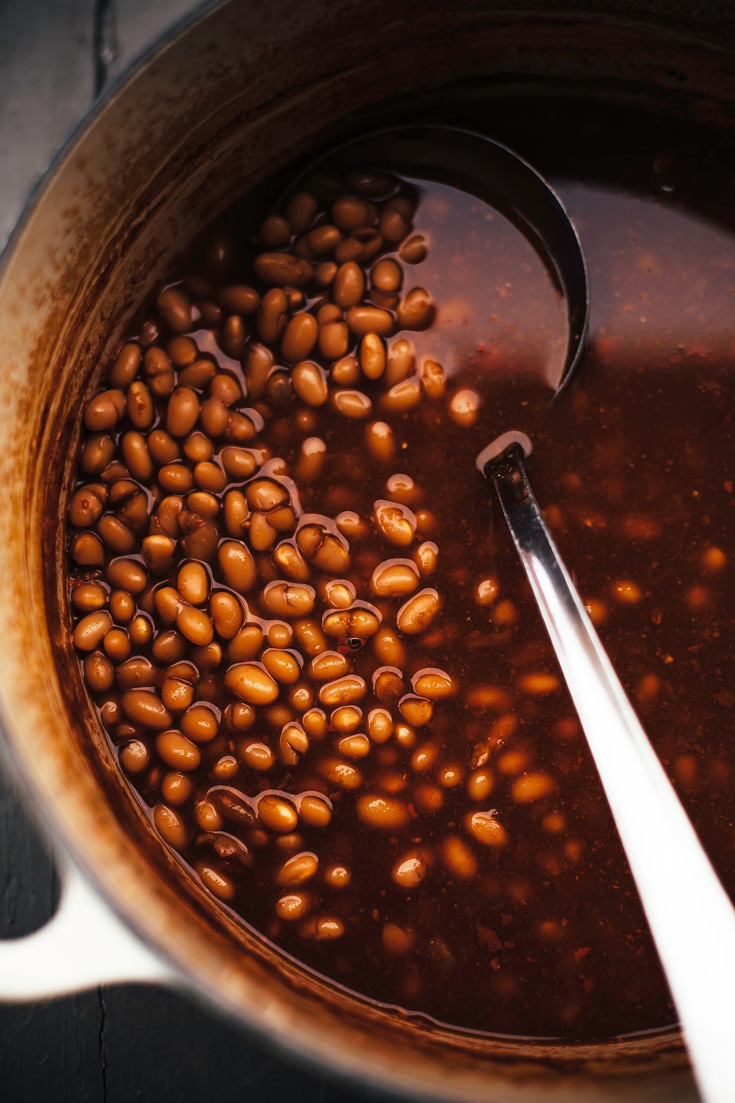 An overhead shot of miso and molasses baked beans in a dutch oven with a ladle inside the pot as well