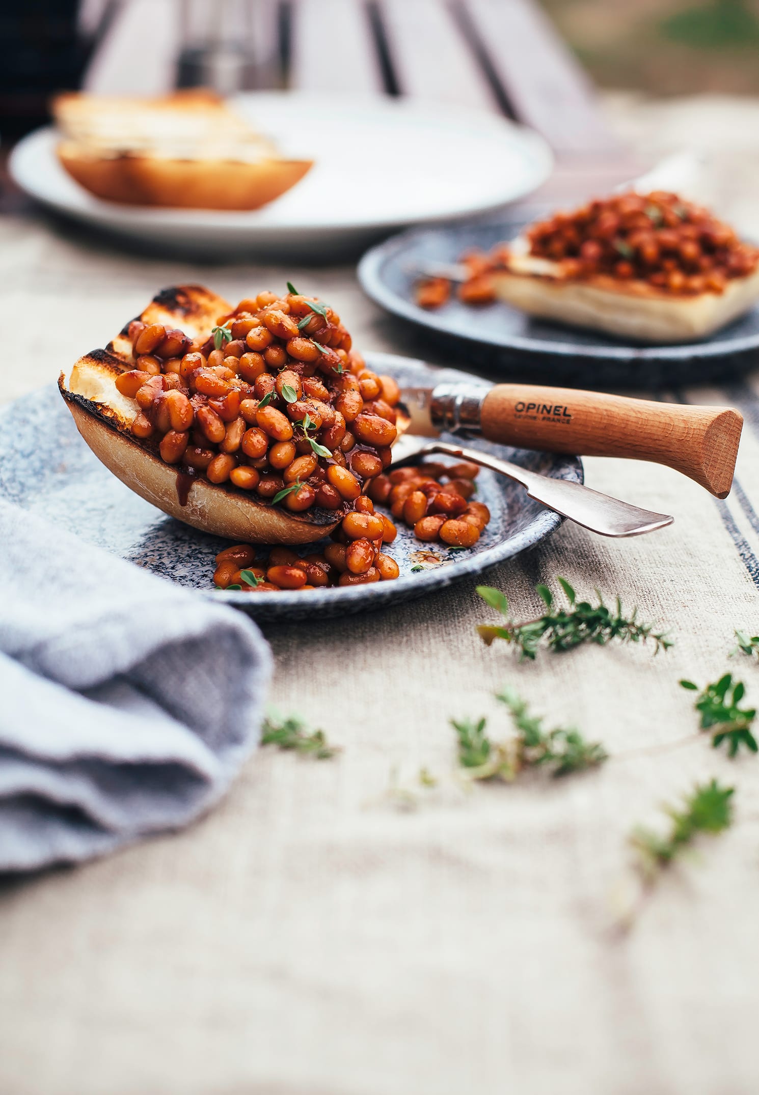 A head on shot of some saucy and brown baked beans piled onto a grilled piece of ciabatta, all on top of a blue enamelware plate.
