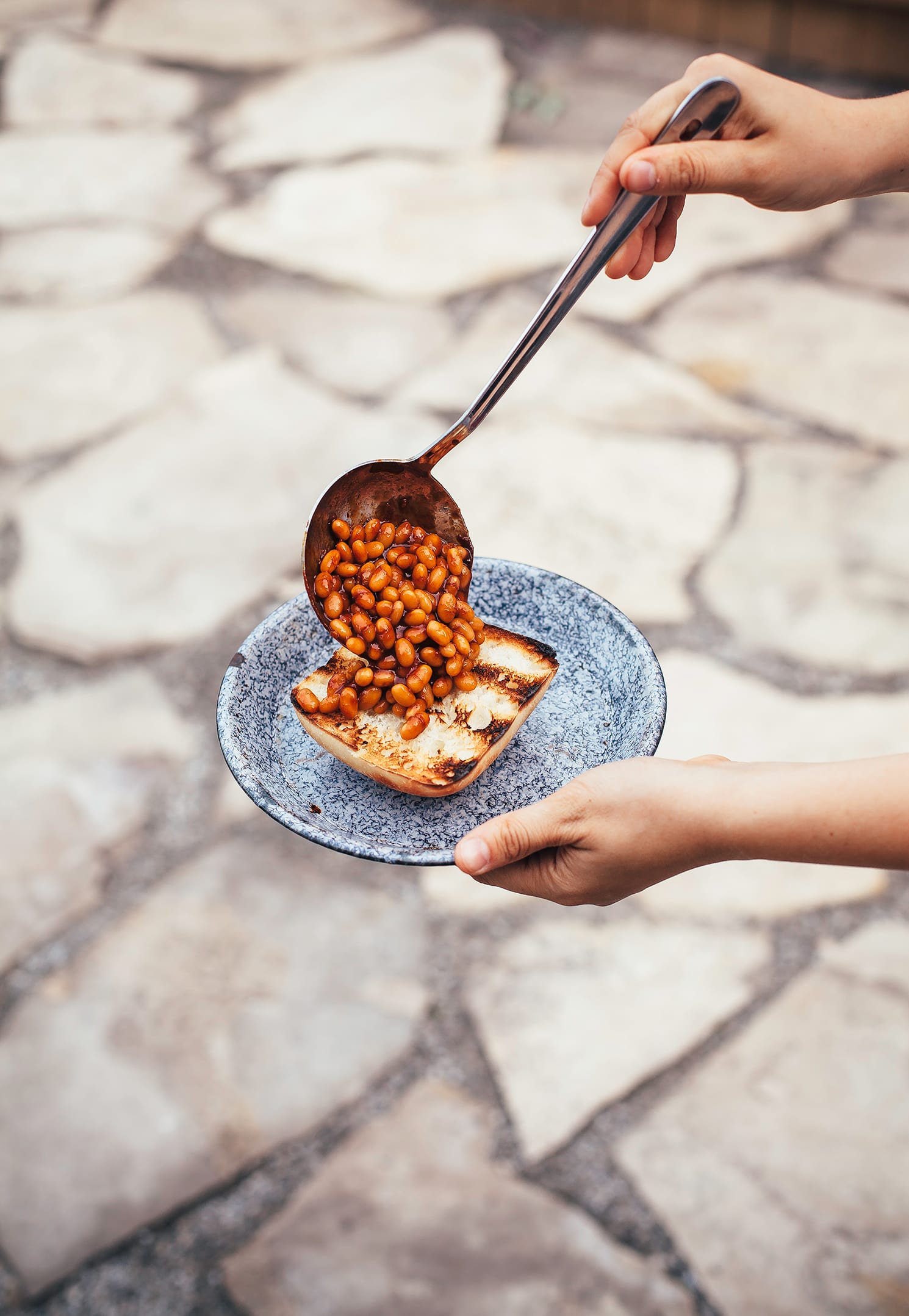 Image shows a hand ladling some brown and saucy baked beans onto a piece of grilled bread.