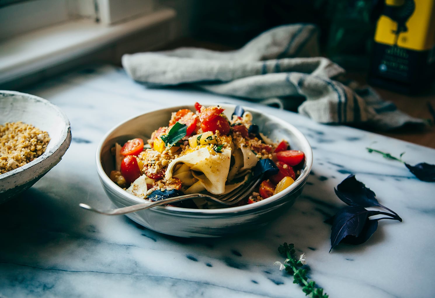 A 3/4 angle shot of a bowl of pasta with chopped tomatoes, fresh basil, and a savoury crumble-like topping.