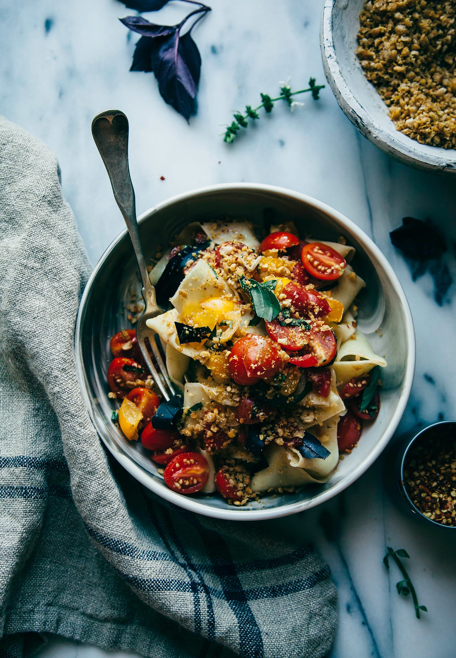 An overhead shot of a bowl of pasta with chopped tomatoes and a savoury crumble-like topping.
