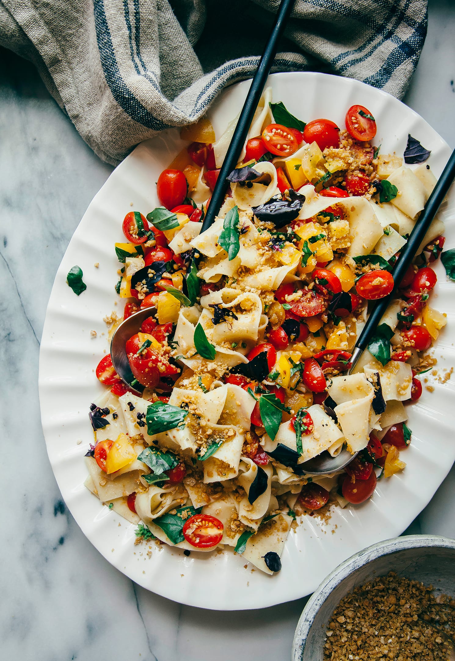 An overhead shot of a platter of pasta with chopped tomatoes, fresh basil, and a savoury crumble-like topping.