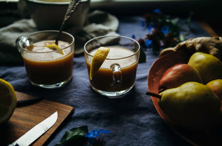 A 3/4 angle shot of warm cider in 2 clear mugs. Pears, ginger root, and a halved lemon are seen to the side.