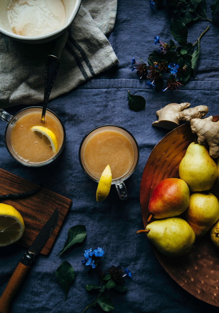 An overhead shot of warm cider in 2 clear mugs. Pears, ginger root, and a halved lemon are seen to the side.